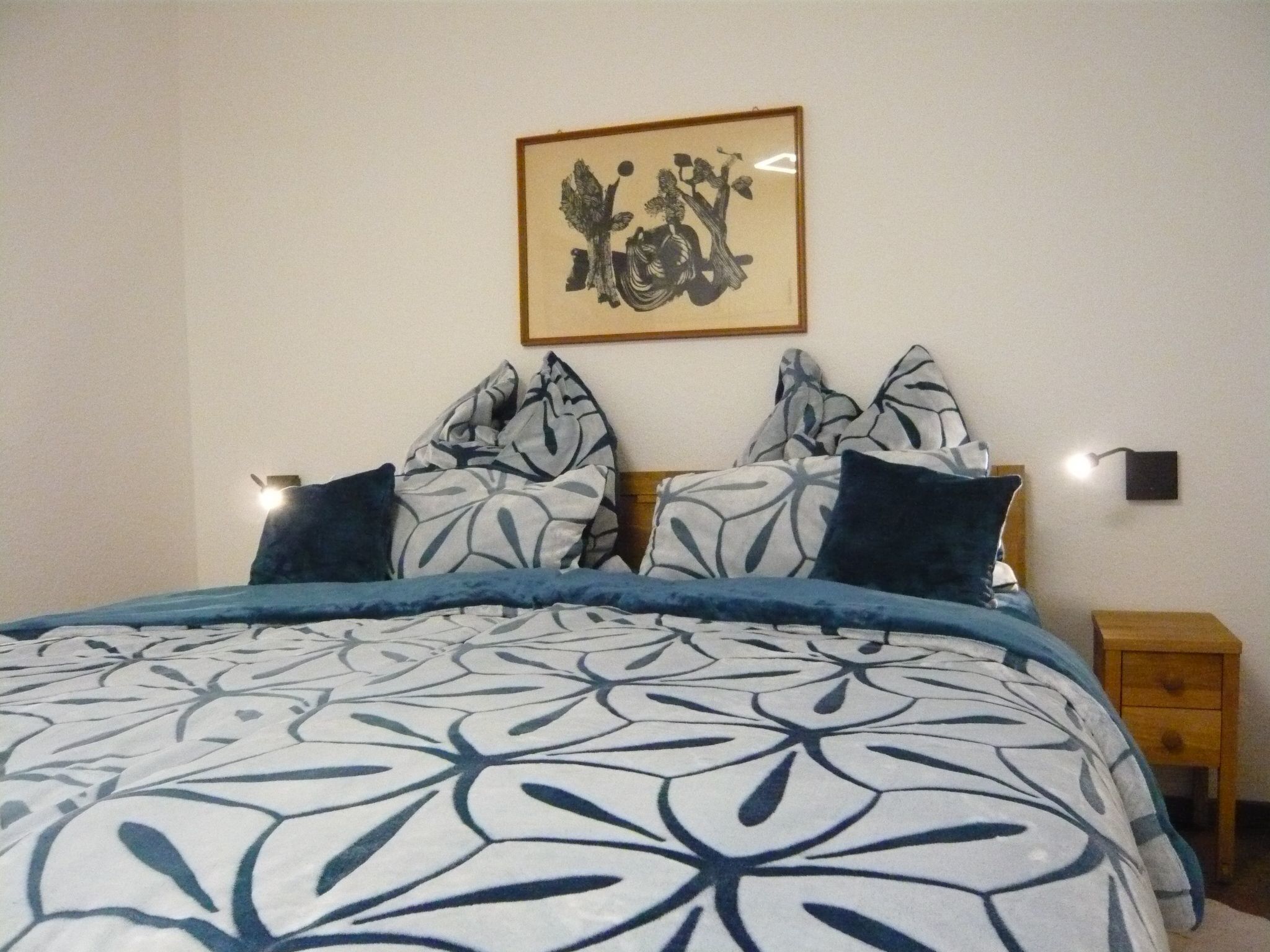 A neatly made bed with a patterned bedspread, a framed artwork above it, and a bedside table with a lamp.
