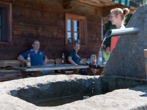 En rustik trästuga med en stenvägg i förgrunden och människor som sitter vid ett träbord.
