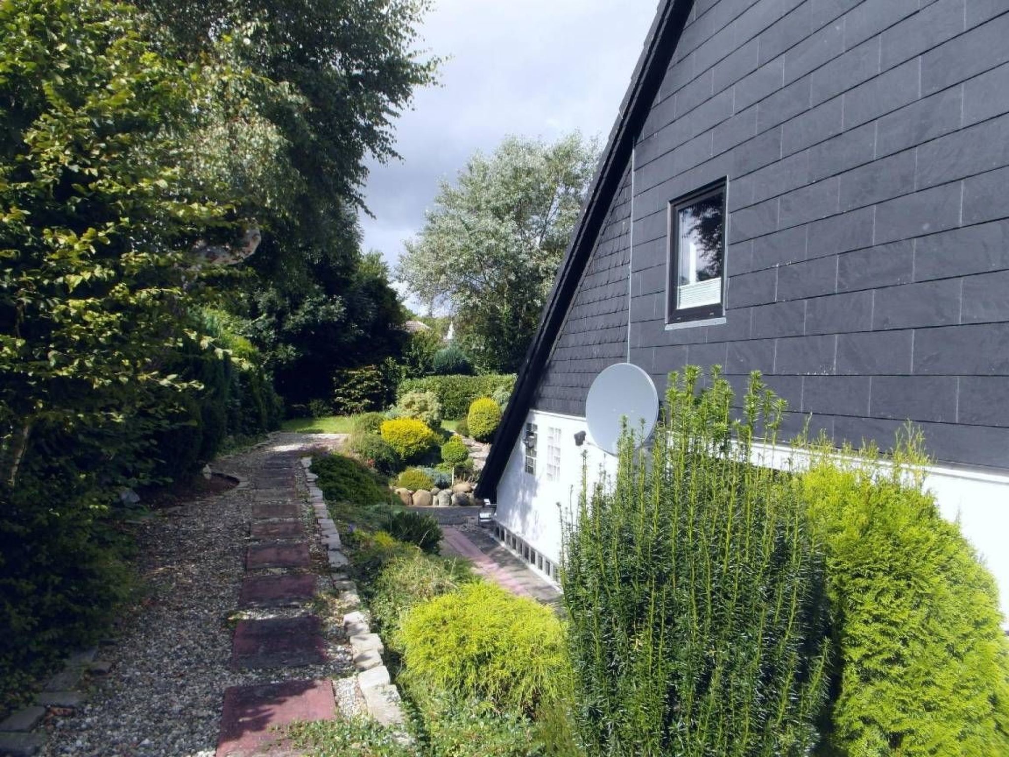 Una casa con facciata scura, un'antenna parabolica visibile e un giardino con diverse piante e un vialetto in ghiaia.