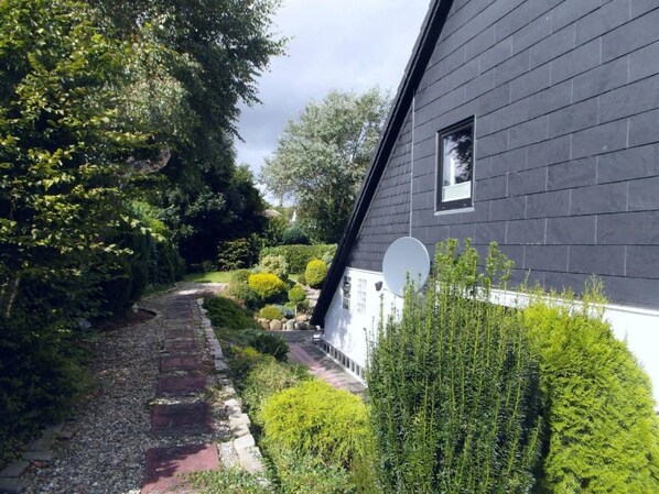 A house with a dark exterior, a visible satellite dish, and a garden with various plants and a gravel path.