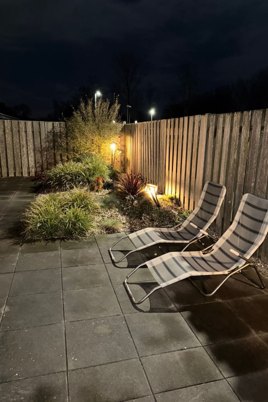 A patio with two lounge chairs, a wooden fence, and a garden area with plants and a lamp post.