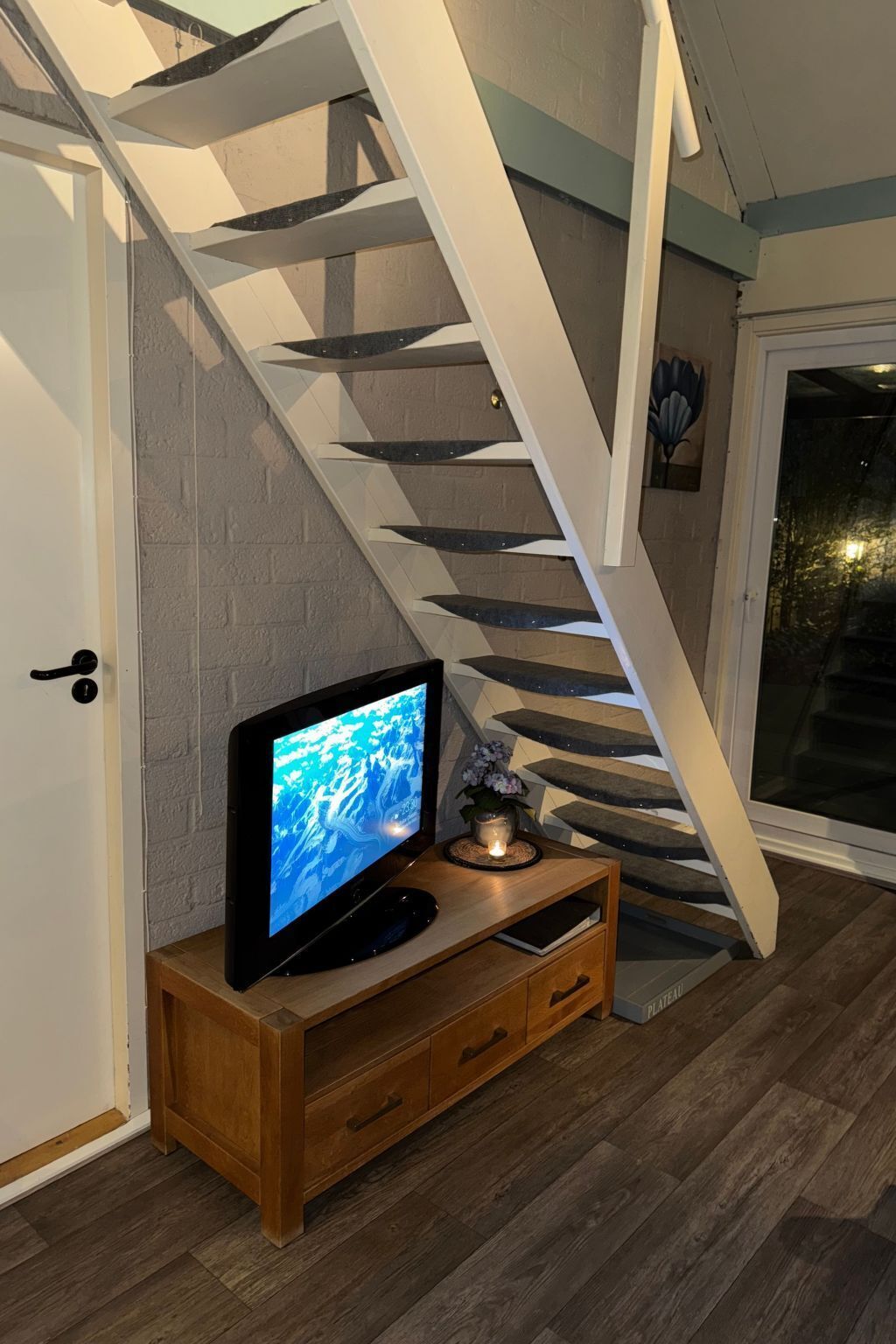 A modern interior with a wooden entertainment unit holding a flat-screen TV, a staircase with a white railing, and a door leading to an outdoor area.