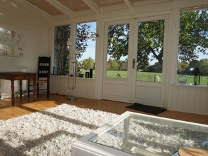 Ein Zimmer mit Holzboden, einem weißen, flauschigen Teppich, einer Glasvitrine, einem Tisch und Stühlen. Große Fenster bieten Blick auf ein grünes Feld und Bäume.