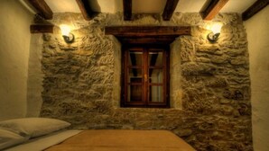 Ein Schlafzimmer mit Steinwand, Holzbalken, einem Fenster mit Holzläden und einem Bett mit weißer Matratze.