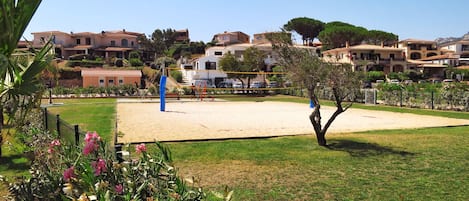 Una cancha de voleibol de arena rodeada por una valla, con un árbol y plantas con flores al frente.