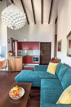 A modern living room with a teal sofa, a wooden coffee table with fruit, a pendant light, and a kitchen in the background. - Residenza Viramonte - Casa Bianca, Wohnung 14 by Interhome (Vira)