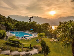 A swimming pool with lounge chairs and umbrellas, surrounded by greenery and mountains in the background. - Residenza Viramonte - Casa Fiona, Wohnung 52 by Interhome (Vira)