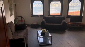 En stue med sofa, sofabord og fjernsyn. Tre vinduer giver udsigt over en sø og bakker.