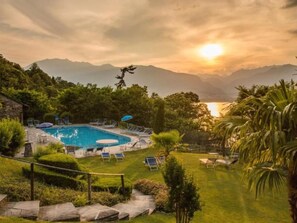 Ein Poolbereich mit Liegestühlen und Sonnenschirmen, umgeben von viel Grün und Bergen im Hintergrund.