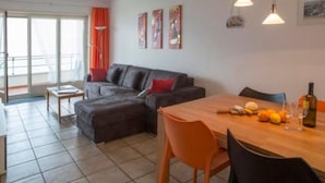 A modern living room with a dark grey sofa, orange cushions, a wooden dining table with chairs, and a balcony with orange curtains. - Residenza Viramonte - Casa Cecilia, Wohnung 24 by Interhome (Vira)