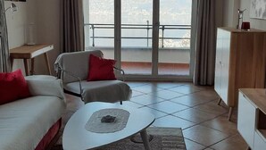 Un salón moderno con sofá, sillón y mesa de centro. Un amplio ventanal corredero da a un balcón con vistas a la ciudad.