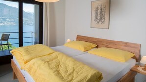 Ein Schlafzimmer mit einem großen Bett, einem hölzernen Kopfteil, einem Nachttisch mit Lampe und einem Fenster mit Blick auf einen See und Berge.