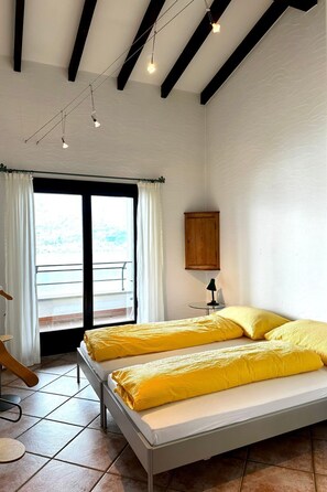 A bedroom with two beds, a wooden cabinet, a small table with a lamp, and a view of the outside through a large window. - Residenza Viramonte - Casa Bianca, Wohnung 19 by Interhome (Vira)
