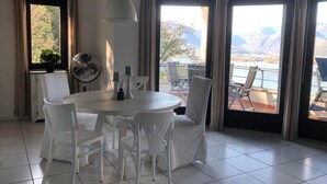 Un comedor con una mesa redonda y sillas blancas, un ventilador y vistas a montañas a través de amplios ventanales.