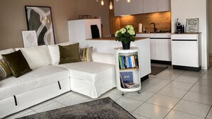 Una sala de estar moderna con un sofá seccional blanco, una pequeña estantería y una cocina al fondo.
