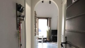 Un pasillo con una puerta de madera oscura, una pared blanca con un estante y vista a una habitación con televisor y planta.