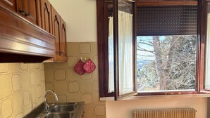 Una cucina con un lavello doppio, mobili impiallacciati in legno, schienale decorato, una finestra con tende e un termosifone.