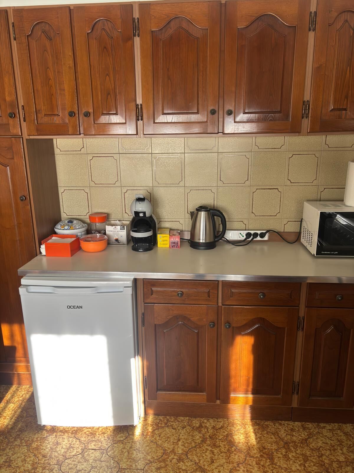 A compact kitchen with wooden cabinets, a refrigerator, a coffee maker, a kettle, and a microwave.