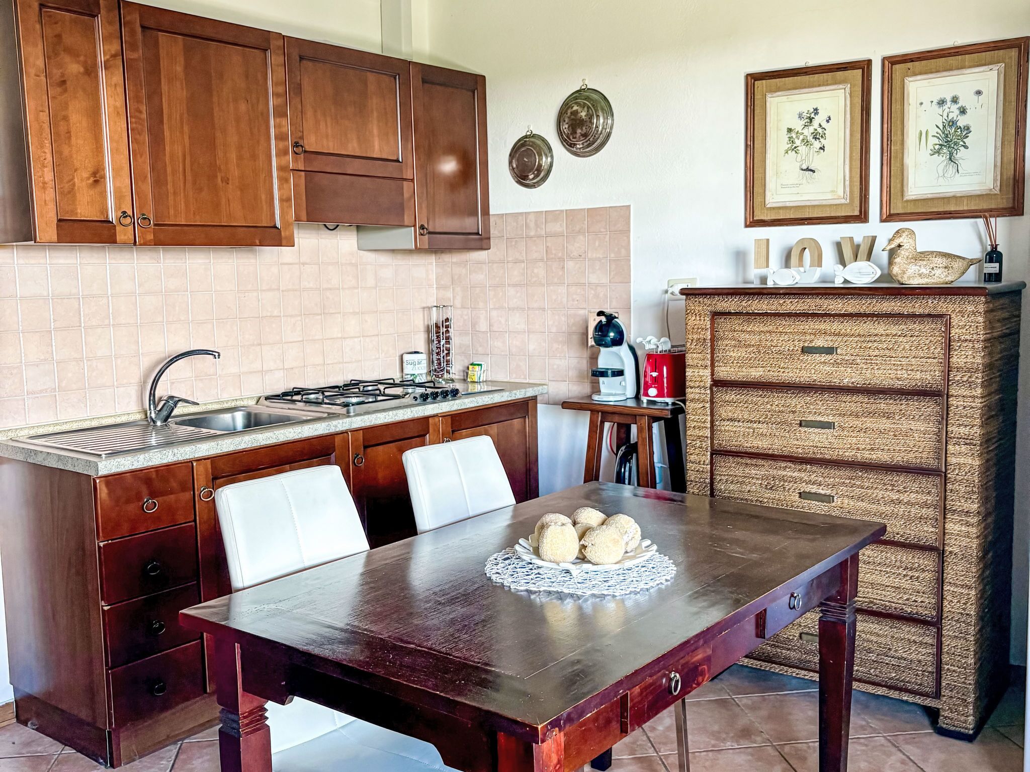 Una cocina con armarios de madera, salpicadero de azulejos, estufa, fregadero, una mesa de comedor con sillas y un aparador de mimbre.