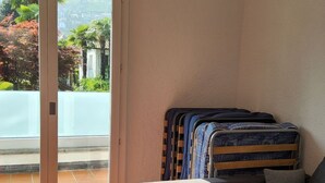 A room with a grey sofa, a bed with a patterned blanket, and a sliding glass door leading to a balcony with a view of greenery and buildings. - Haus in Agno by Interhome (Agno)