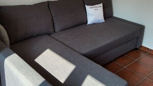 A grey sectional sofa with a white pillow against a plain wall, positioned on a tiled floor. - Haus in Agno by Interhome (Agno)
