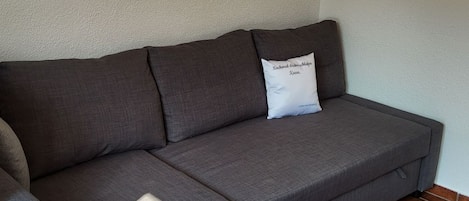 A grey sectional sofa with a white pillow against a plain wall, positioned on a tiled floor.