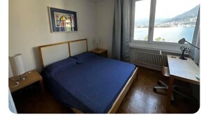 A bedroom with a bed, a desk, a chair, a radiator, and a window with a view of a body of water and mountains. - Paradise Lake View by Interhome (Lugano)
