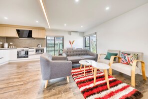 A modern living space with a kitchen, grey sofas, a white coffee table, and a red and white striped rug. - Cosy & Contemporary Airport Retreat (Papatoetoe Auckland)