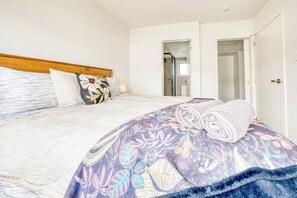 A bedroom with a bed, a wooden headboard, a floral-patterned bedspread, a rolled towel, and a door leading to a bathroom. - Cosy & Contemporary Airport Retreat (Papatoetoe Auckland)