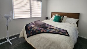 A bedroom with a bed, a standing fan, a window with blinds, and a ceiling light. - Cosy & Contemporary Airport Retreat (Papatoetoe Auckland)