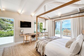 Ein Schlafzimmer mit einem großen Bett, einem Schreibtisch mit Stuhl, einem an der Wand montierten Fernseher und einem Balkon mit Meerblick.