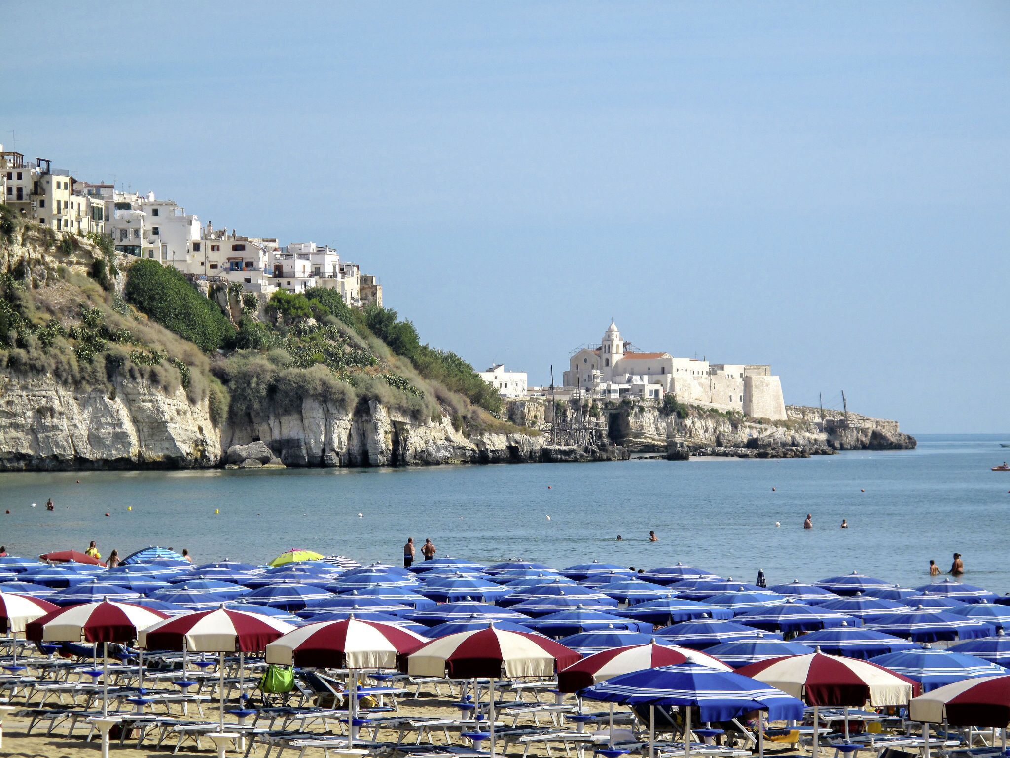 Ein Strand mit vielen Sonnenschirmen und Liegestühlen, eine Küstenstadt mit Gebäuden auf einer Klippe und das Meer im Hintergrund.