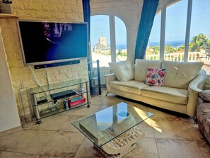 A living room with a flat-screen TV mounted on a textured wall, a beige sofa with a floral pillow, a glass coffee table, and a view of the sea through large windows. - Casa Flamingo by Interhome (Calpe/Calp)