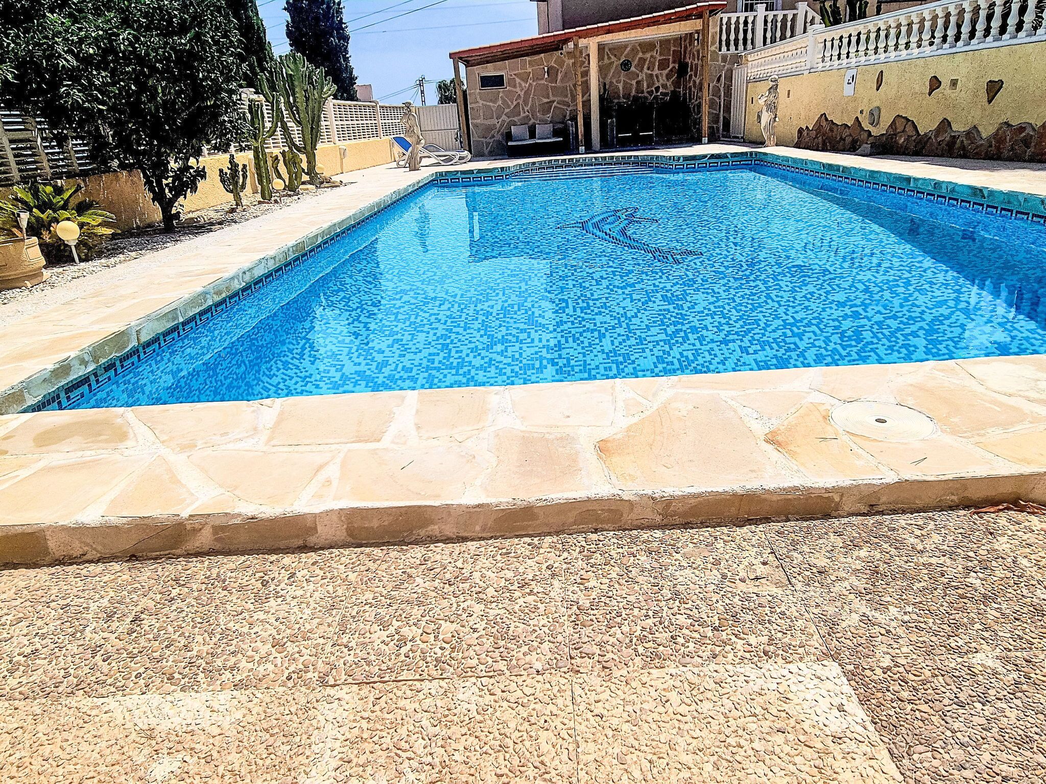 A swimming pool with a stone border, surrounded by a patio area with seating and a building in the background.