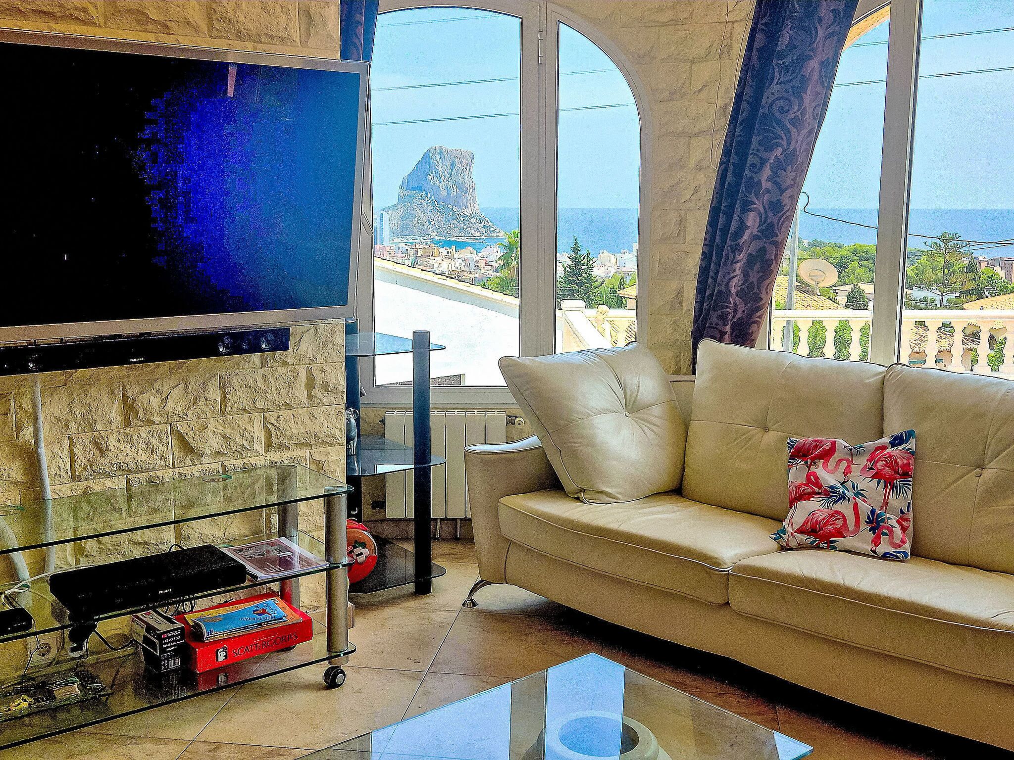 A living room with a sofa, a flat-screen TV mounted on a stone wall, a glass coffee table, and a view of the sea and a mountain.