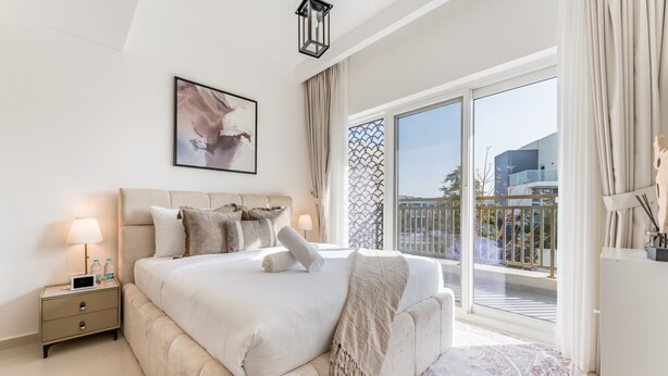 Une chambre moderne avec un grand lit, une table de chevet avec une lampe, un tableau encadré au mur et une vue sur le balcon extérieur.