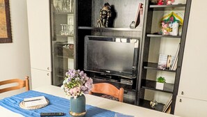 Un comedor con una mesa, sillas, un florero con flores, un control remoto y un televisor.