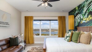 A bedroom with a bed, a sofa, a window with curtains, a ceiling fan, and a view of a body of water. - Lazy River - Lake Views - Hot Tub - Pet Frndly! (Kimberling)