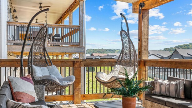 Una terraza de madera con mobiliario de mimbre, incluyendo sillones colgantes y un sofá, con vista a un paisaje verde.