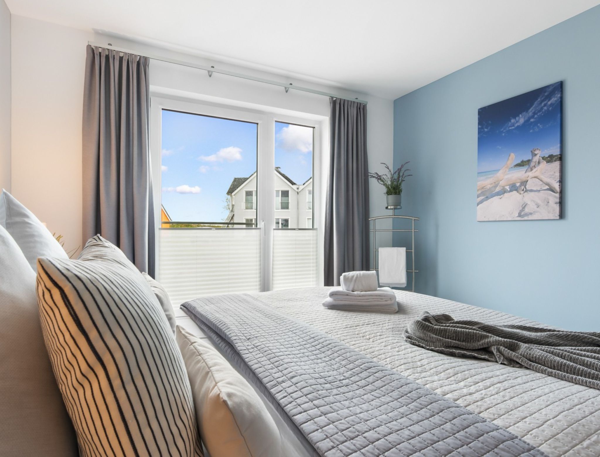 Een slaapkamer met een bed, kussens, een nachtkastje met een plant, een handdoek en een schilderij van een strand.