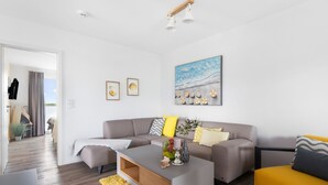 A modern living room with a grey sofa, a yellow armchair, and a coffee table. There are framed pictures on the wall and a painting above the sofa. - Vacation apartment "Dünengras" in the Baltic Sea resort of Olpenitz - Modern accommodation with a street (Kappeln)