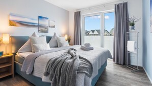 A modern bedroom with a large bed, a bedside table, a nightstand, a window with curtains, and a view of houses outside. - Vacation apartment "Dünengras" in the Baltic Sea resort of Olpenitz - Modern accommodation with a street (Kappeln)