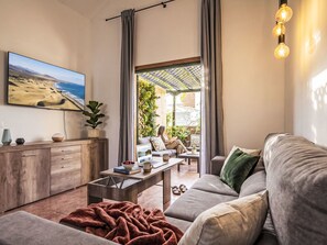 Ein modernes Wohnzimmer mit einer Couch, einem Couchtisch und einem Fernseher. Es gibt einen Blick auf einen Balkon mit Pflanzen und einer draußen sitzenden Person.