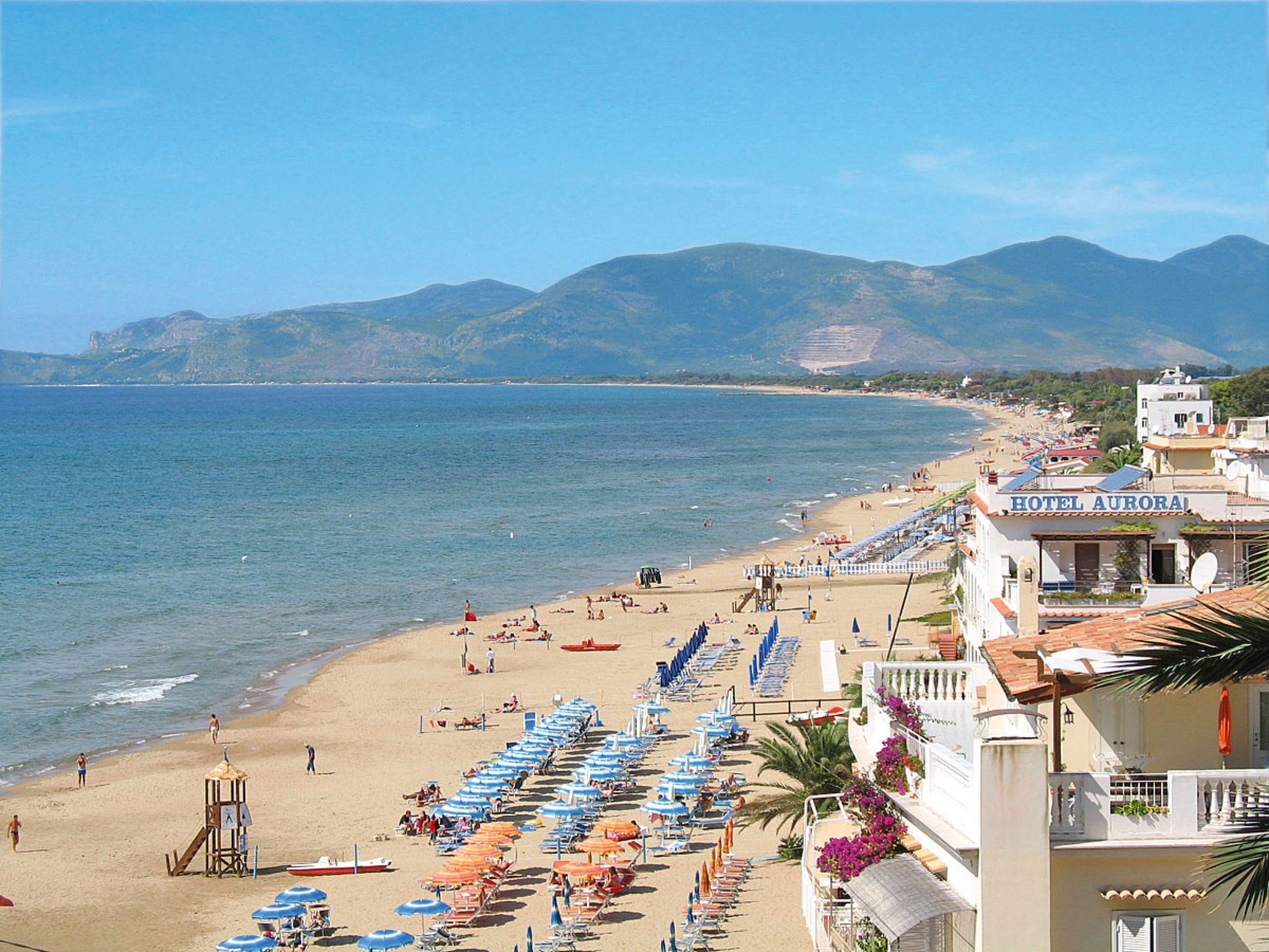 A beach with rows of sunbeds and umbrellas, a lifeguard tower, and a hotel named 'HOTEL AURORA' with a view of the sea and mountains.