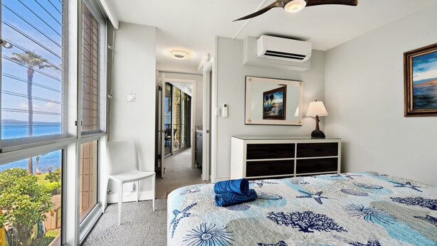A bedroom with a large window, a ceiling fan, a bed with a blue and white patterned bedspread, a white dresser, and a painting on the wall. - Kamaole Nalu 403 - Ocean View - New DESIGNER Remodel wA/C (Kihei)