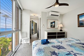 A bedroom with a large window, a ceiling fan, a bed with a blue and white patterned bedspread, a white dresser, and a painting on the wall. - Kamaole Nalu 403 - Ocean View - New DESIGNER Remodel wA/C (Kihei)
