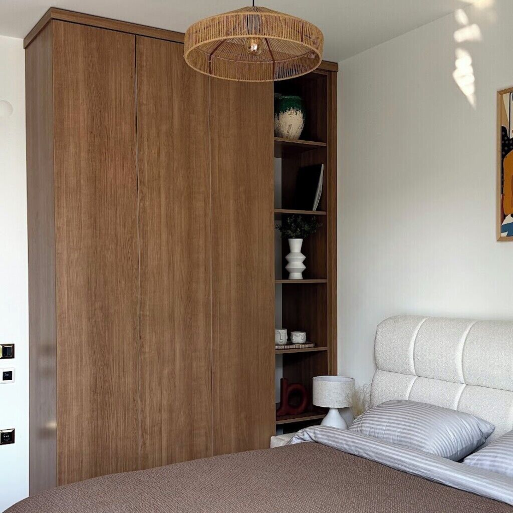 A bedroom with a large wooden wardrobe, a bed with a headboard, a bedside table, and a hanging light fixture.