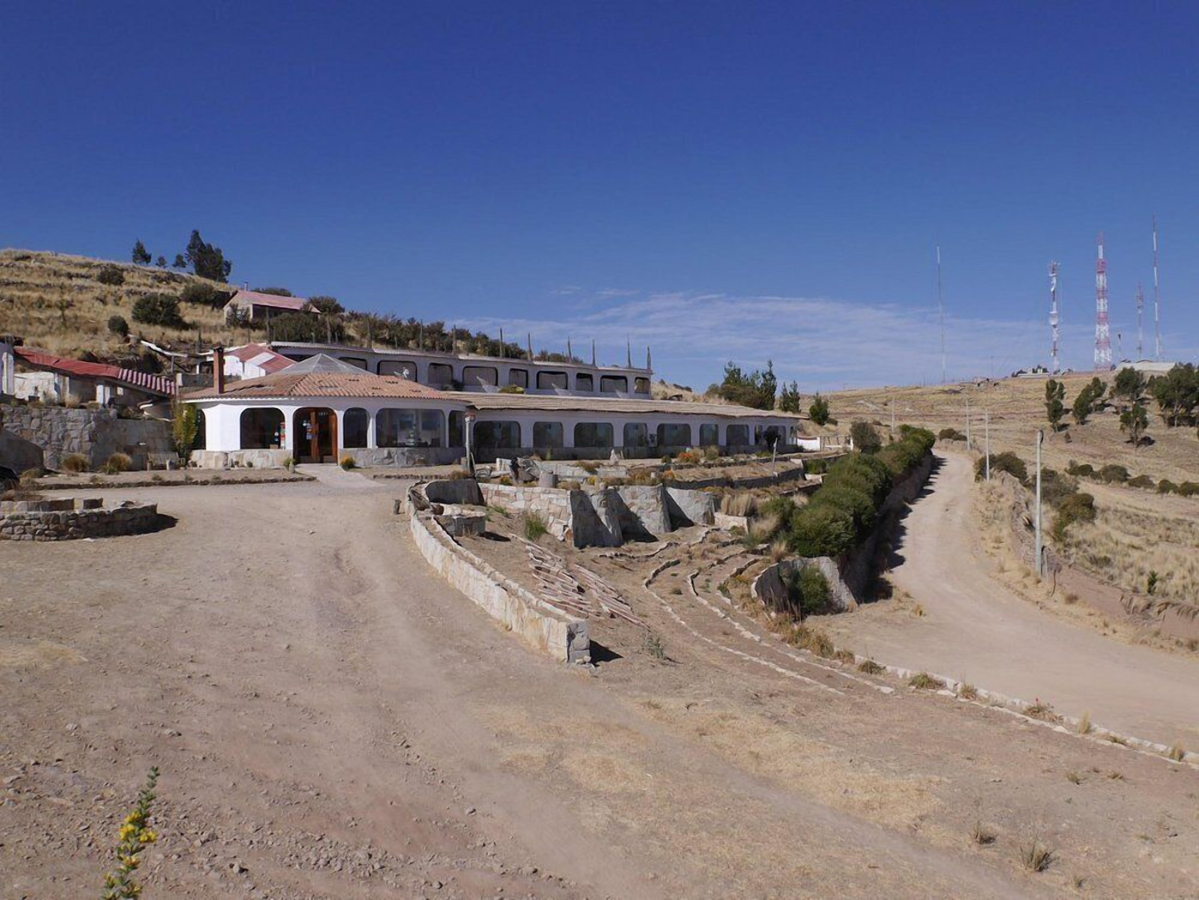 A large, white building with multiple windows and a red-tiled roof, surrounded by a dry, hilly landscape.