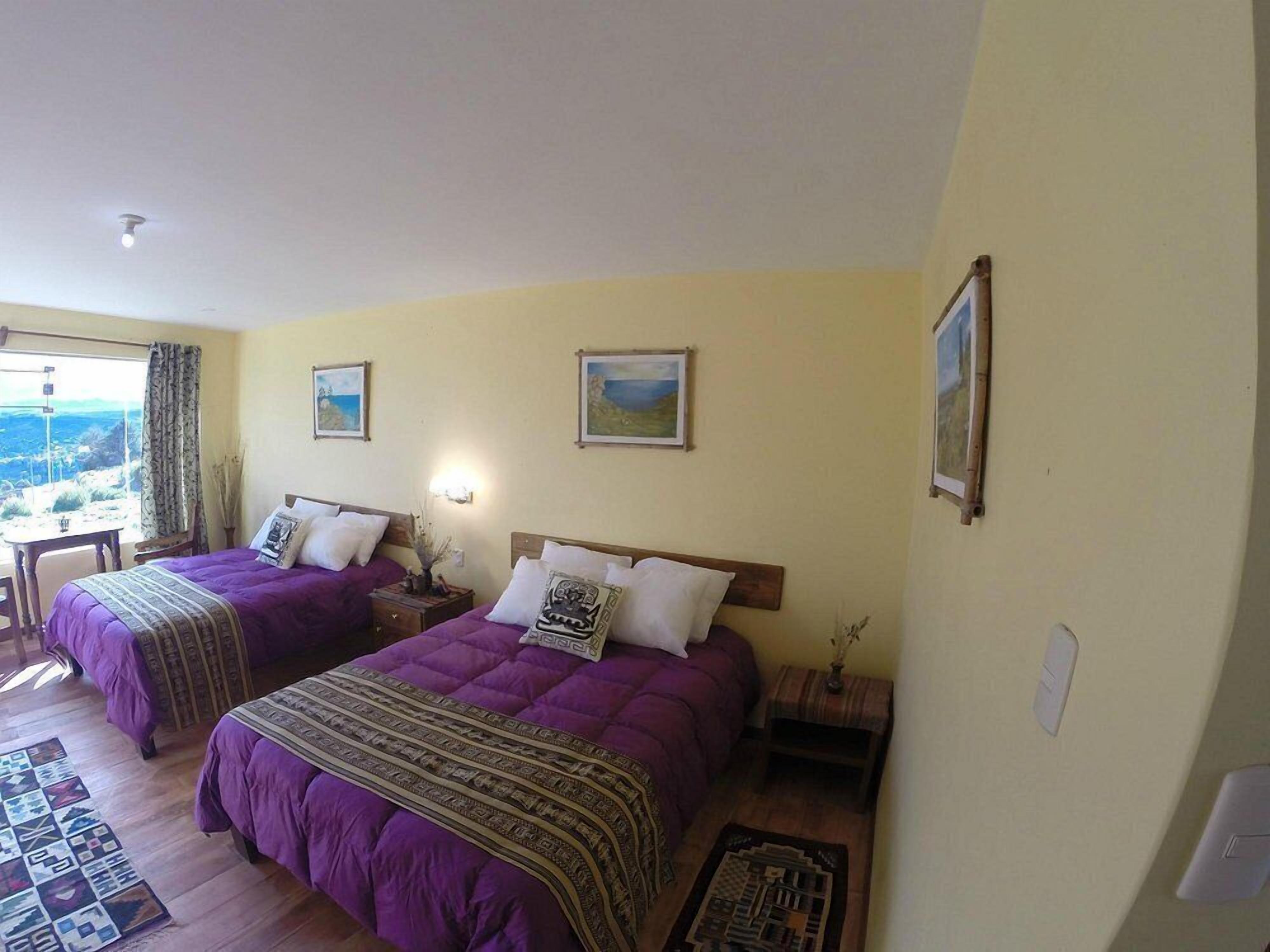 A room with two beds, each with purple bedding and pillows, a small table, and a chair. There are framed pictures on the walls and a window with a view of the outdoors.