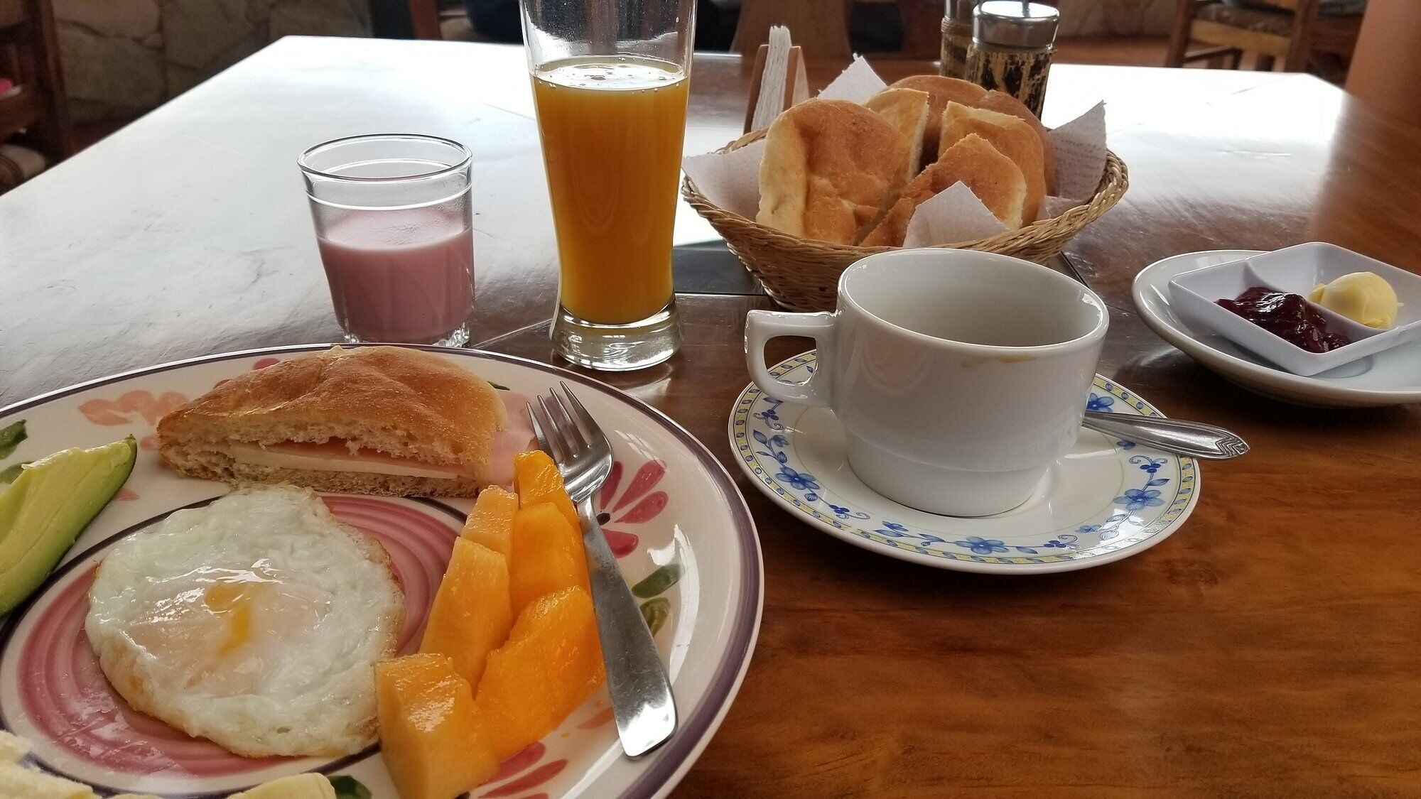 Mesa de café da manhã com sanduíche, ovo, frutas, copo de suco de laranja, xícara de café e cesta de pão.